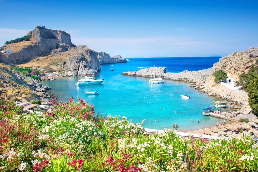 Panoramablick auf die türkisblaue St. Pauls-Bucht bei Lindos auf Rhodos mit weißen Segelbooten, umrahmt von blühenden Pflanzen im Vordergrund und der antiken Akropolis auf dem Felsen im Hintergrund – ein traumhaftes Ziel für Last Minute Griechenland Urlauber.