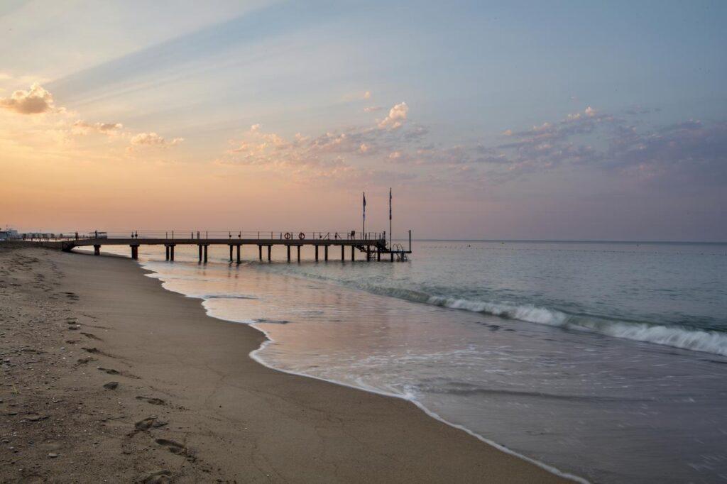 Sandstrand mit Holzsteg führt ins ruhige Meer bei Sonnenuntergang, pastellfarbener Himmel in Rosa und Orange, sanfte Wellen am Strand des TUI MAGIC LIFE Belek