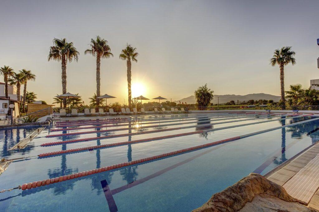 Lastminute Sommerurlaub: Olympisches Sportbecken mit Bahnen im TUI MAGIC LIFE Marmari Palace bei Sonnenuntergang, umgeben von Palmen und mit Meerblick auf Kos
