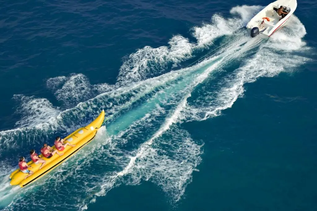 Luftaufnahme von gelbem Bananenboot mit mehreren Personen und weißem Motorboot, die Kurven durch tiefblaues Meer ziehen und weiße Gischtspuren hinterlassen