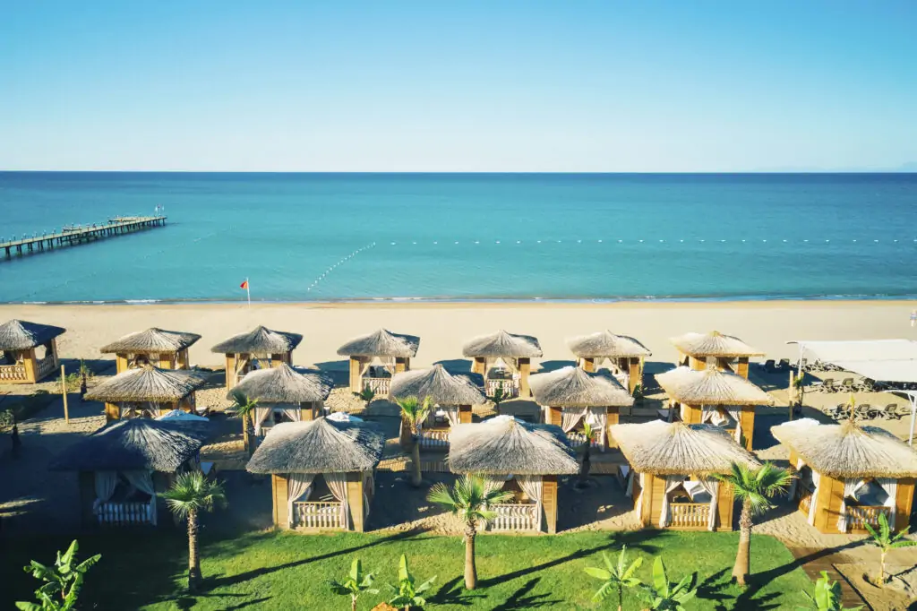 Strandabschnitt des TUI MAGIC LIFE Masmavi mit Reihen von strohgedeckten Beach-Cabanas auf grüner Wiese, weißem Sandstrand, türkisblauem Meer und Holzsteg im Hintergrund