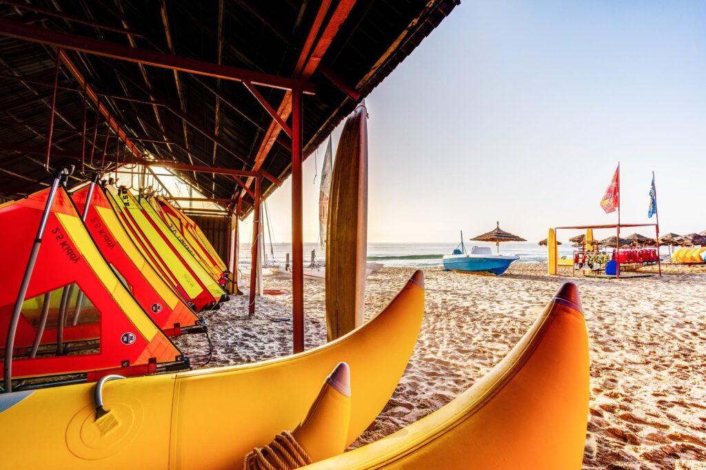 Wassersport-Station am Sandstrand des TUI MAGIC LIFE Africana mit bunten Kajaks in Orange und Gelb, überdachtem Bereich und Segelbooten im Hintergrund am Mittelmeer