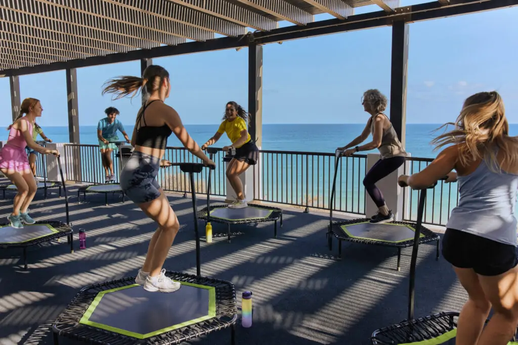 Gruppe von Frauen beim Trampolin-Fitness-Kurs auf kleinen Trampolinen auf einer überdachten Dachterrasse mit Panoramablick auf das türkisblaue Meer im Hintergrund