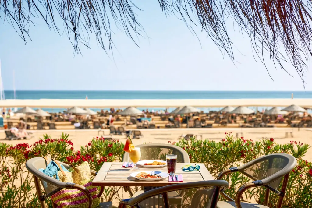 Gedeckter Tisch mit Frühstück und Cocktails auf einer Terrasse mit Meerblick, im Hintergrund Sandstrand mit Sonnenschirmen, grüne Pflanzen im Vordergrund und Pinienzweige über dem Tisch