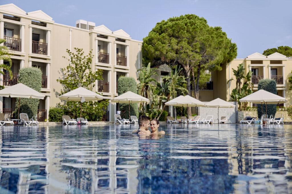 Poolbereich des TUI MAGIC LIFE Belek mit blauem Infinity-Pool, weißen Sonnenschirmen, modernen weißen Hotelgebäuden, Pinien und Menschen im Pool
