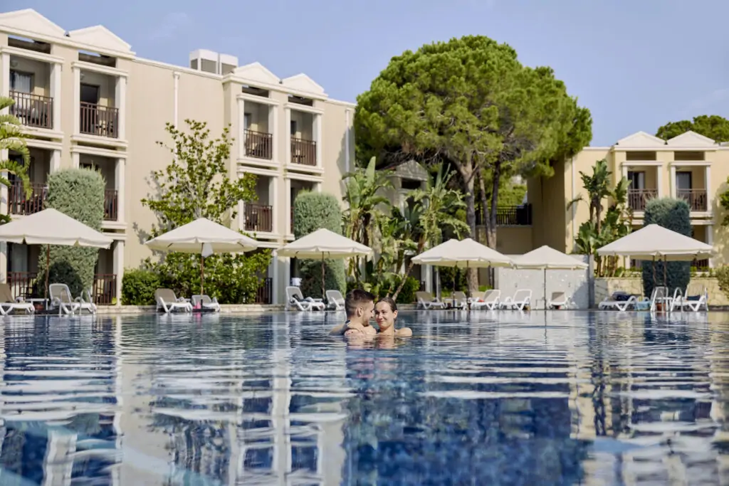 Poolbereich des TUI MAGIC LIFE Belek mit blauem Infinity-Pool, weißen Sonnenschirmen, modernen weißen Hotelgebäuden, Pinien und Menschen im Pool
