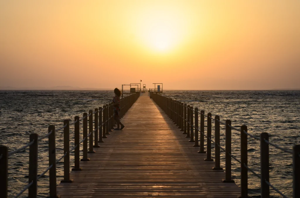 Langer Holzsteg führt bei goldenem Sonnenuntergang ins Rote Meer, Person steht am Ende des Stegs, orange-goldener Himmel spiegelt sich im Wasser