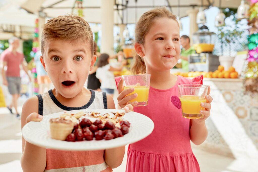Zwei begeisterte Kinder im Buffetbereich eines TUI MAGIC LIFE Clubs. Ein Junge präsentiert freudig überrascht einen Teller mit Obstkuchen und Früchten, während ein Mädchen im rosa Kleid zwei Gläser Orangensaft hält. Im Hintergrund ist das bunte Buffet mit frischen Früchten zu sehen – ein typisches Beispiel für die kinderfreundliche Verpflegung im Last Minute Familienurlaub.