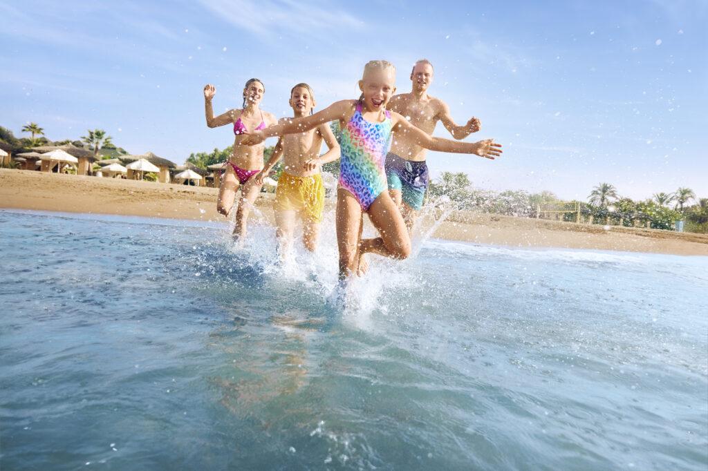 Familie springt gemeinsam lachend ins Meer und erlebt ihren Last Minute Familienurlaub am sonnigen Sandstrand.