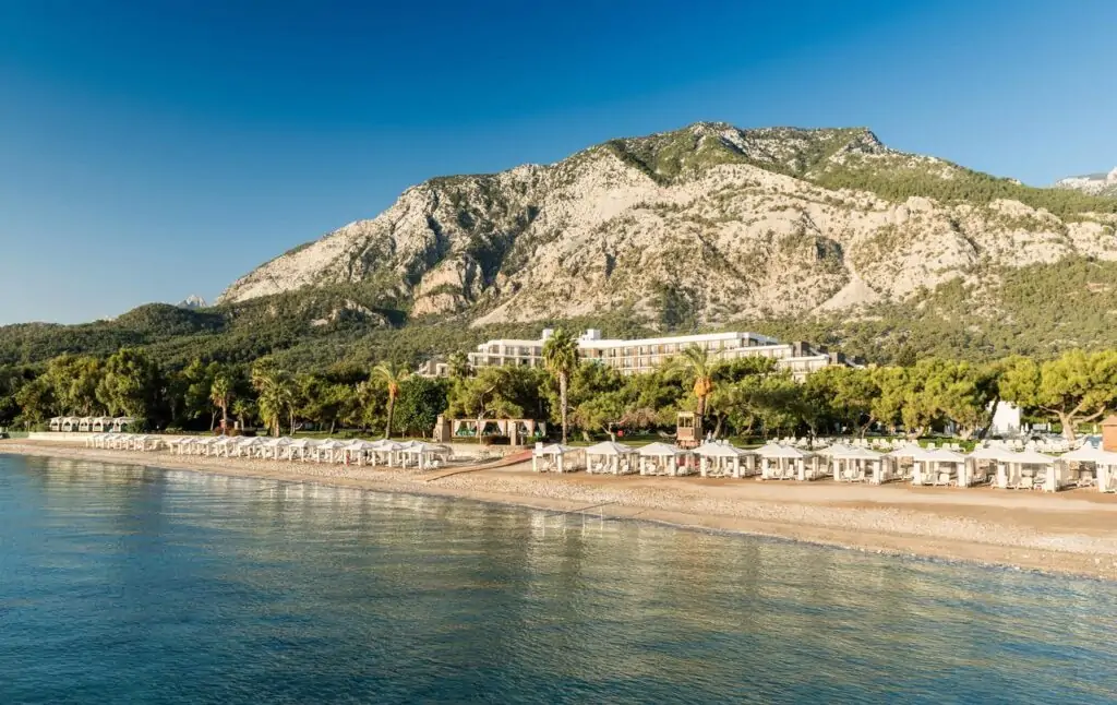 Strandansicht des TUI MAGIC LIFE Beldibi mit weißem Kiesstrand, Reihen von Sonnenschirmen und Liegen, weißem Hotelgebäude zwischen grünen Pinienwäldern und dramatischem Taurusgebirge im Hintergrund