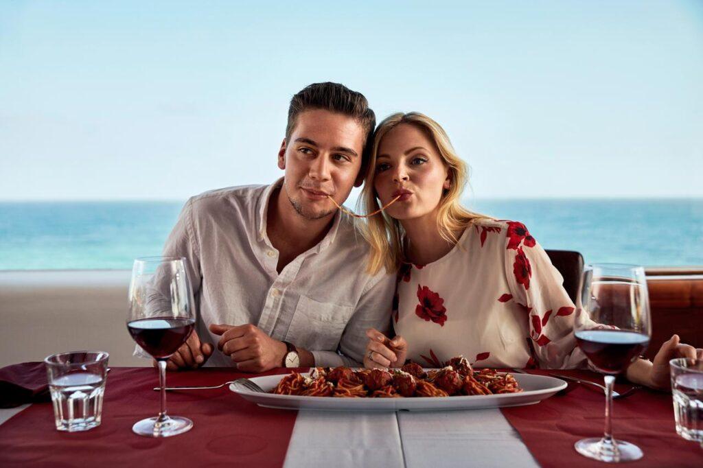 Paar beim romantischen Dinner am Meer während Paarurlaub All Inclusive - Mann und Frau stoßen mit Rotwein an vor mediterranem Meerespanorama