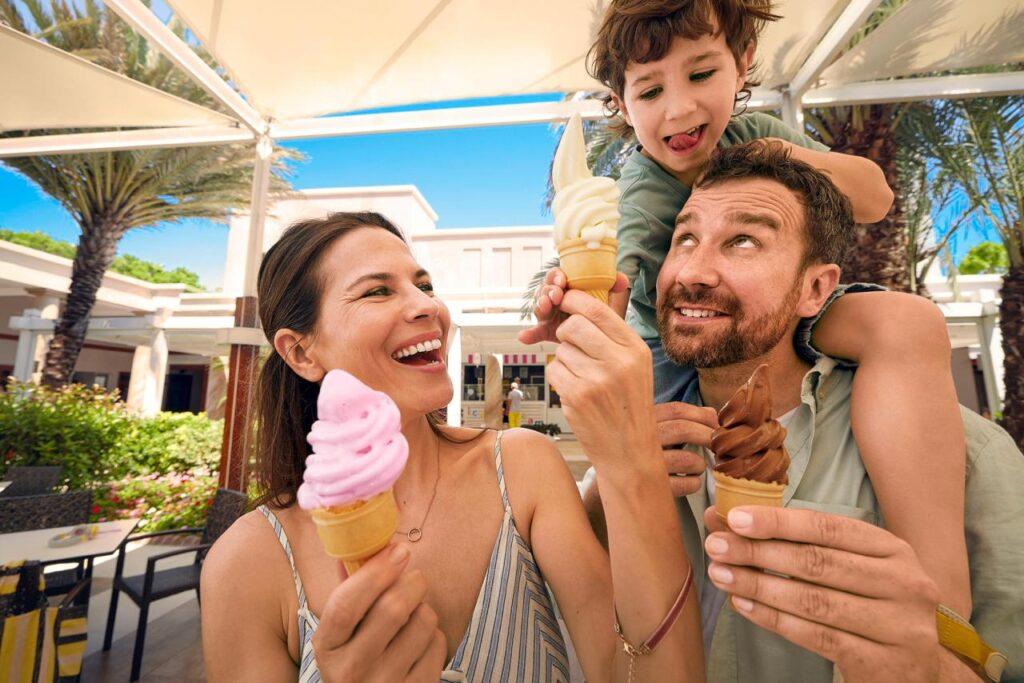 Lachende Familie mit Kleinkind und buntem Eis auf Außenterrasse eines All Inclusive Familienhotels mit Pool