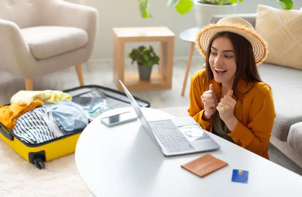 Frau sitzt mit Laptop und Reiseunterlagen auf dem Sofa und plant mithilfe digitaler Tools eine bevorstehende Urlaubreise.