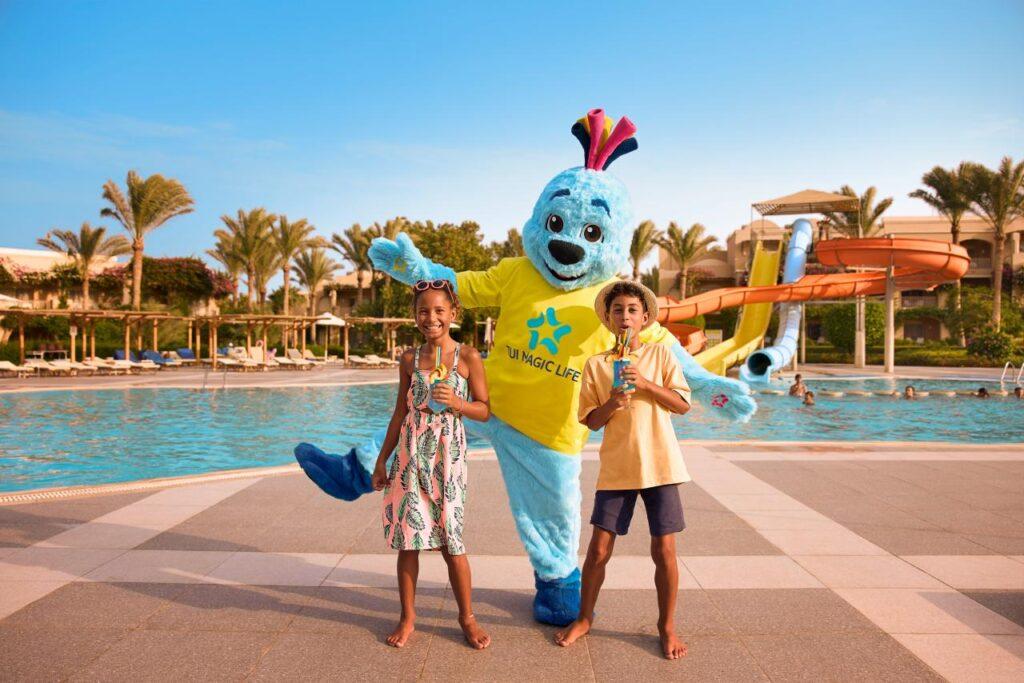 Zwei Kinder mit TUI MAGIC LIFE Maskottchen vor Aquapark mit Wasserrutschen im Hintergrund