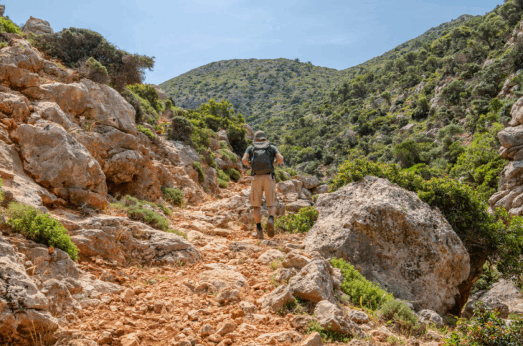 Steiniger Wanderweg in den Bergen von Kreta, umgeben von großen Felsen und mediterraner Vegetation, mit einer Person auf dem Pfad unter klarem Himmel