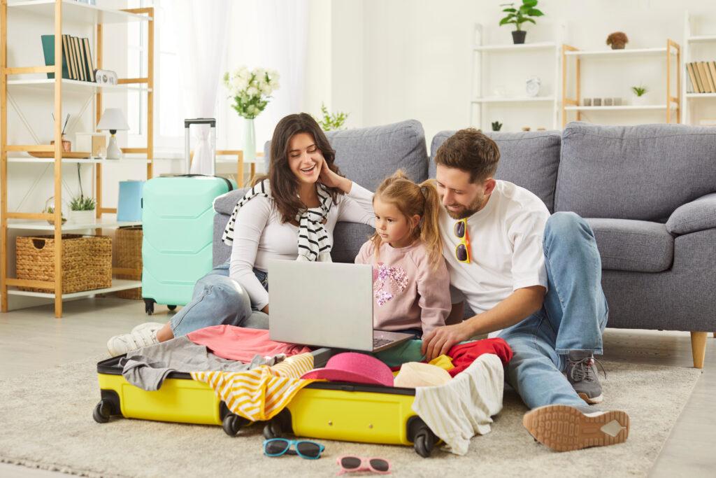 Familie sitzt im Wohnzimmer vor einem offenen Koffer und nutzt einen Laptop zur Reiseplanung – Vorbereitung für Familienurlaub in Kalabrien.